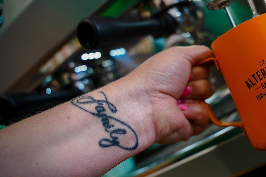 A woman holding a milk frothing jug, the word 'family' tattooed on her wrist.