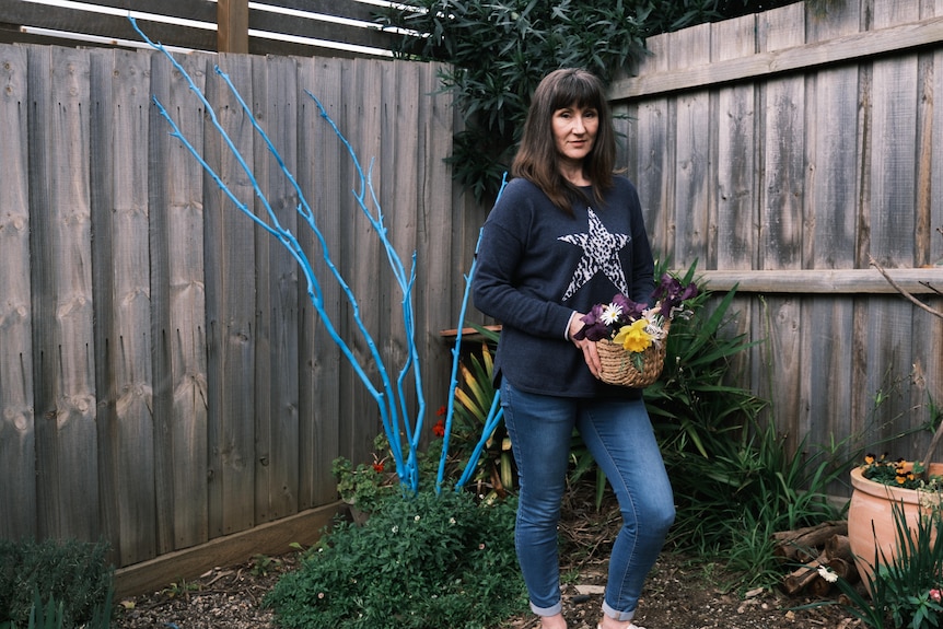 Kim stands with a basket of flowers in front of old tree branches that are painted blue and arranged in a bouquet in the ground.