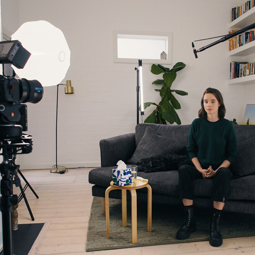 A woman in a black outfit sits on a grey couch in front of a camera and lights