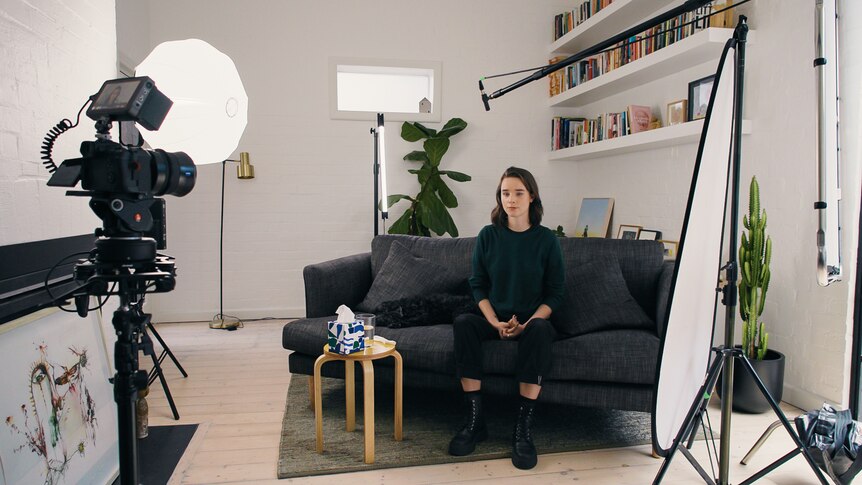 A woman in a black outfit sits on a grey couch in front of a camera and lights