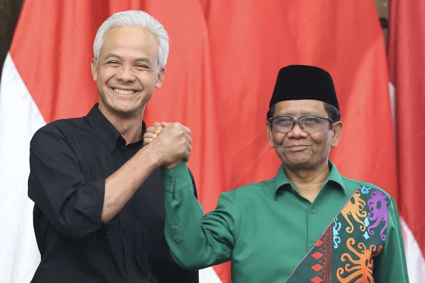A middle-aged man with white hair in a black shirt, together with a man in a black cap and green shirt, smiles at the camera.