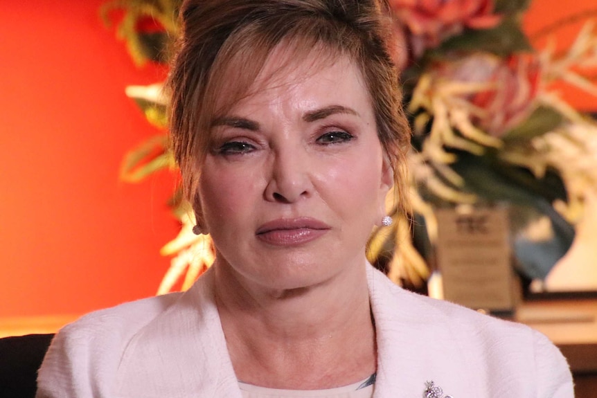 A woman in a pale pink jacket in front of a red background.