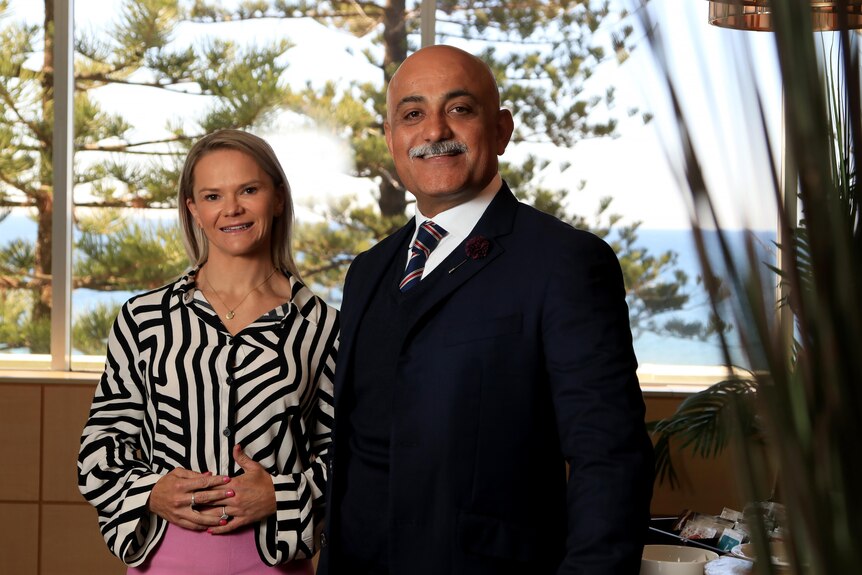 Emily wears a black and white blouse and Ajay wears a three-piece suit. Both are smiling with a window behind them.