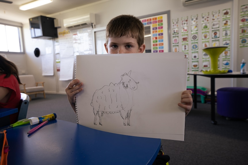 A young boy student holds up a drawing pad marked with a sheep illustration in a classroom. 
