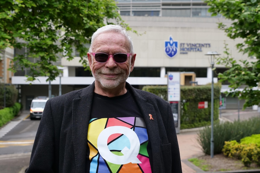 A man wearing pink sunglasses smiles outside a hospital