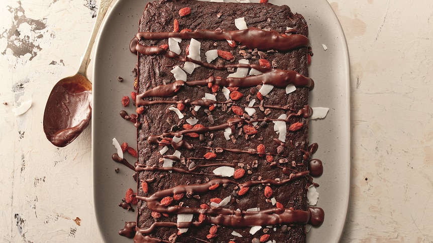 A sweet potato brownie drizzled in chocolate and flakes, alongside a chocolate-covered spoon.