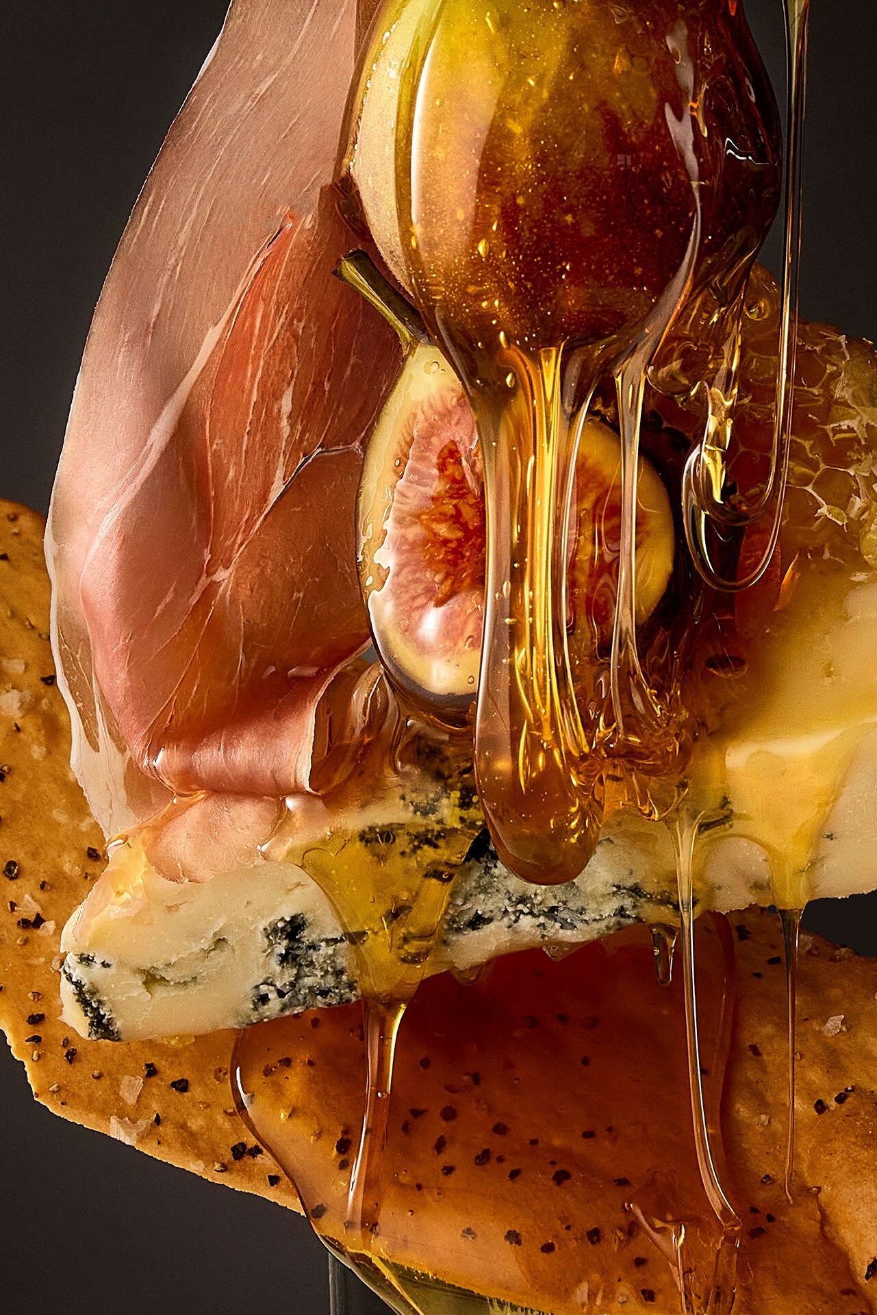 A close-up shot of honey pouring over figs, prosciutto, blue cheese and a cracker.  