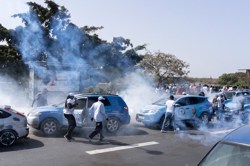 Young men and women cover their faces as tear gas spreads through a crowd of people and cars.
