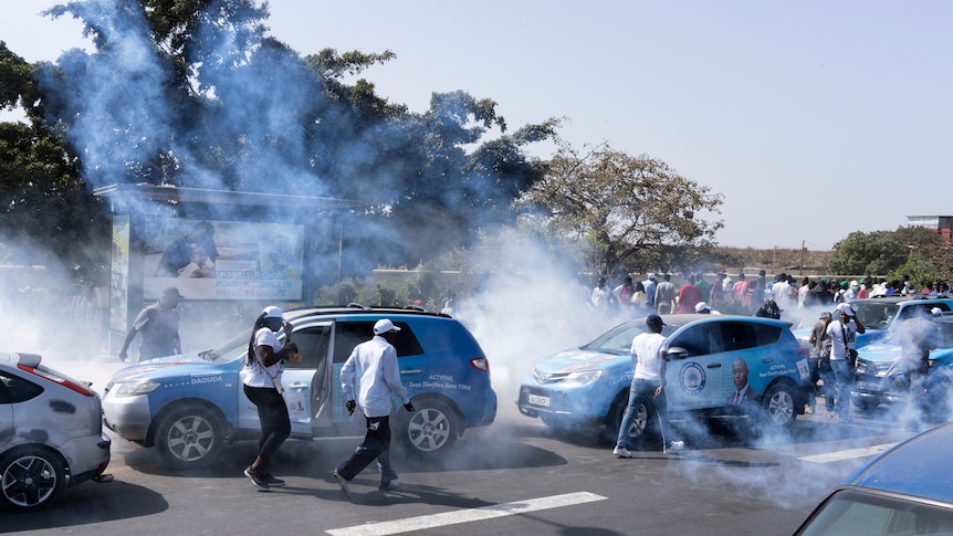 Young men and women cover their faces as tear gas spreads through a crowd of people and cars.