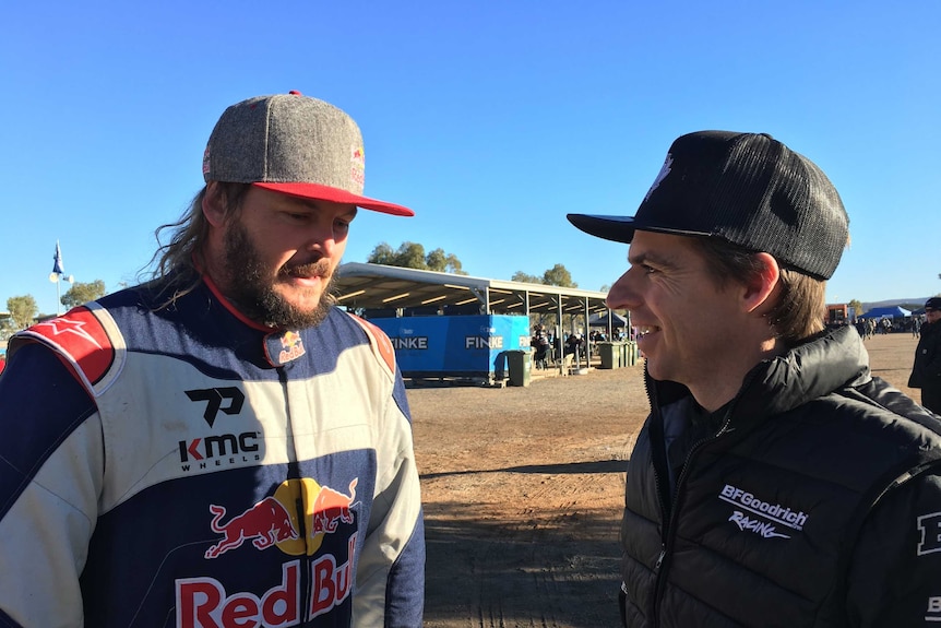 Two men in motorcycle gear and caps stand talking with dirt in the background 
