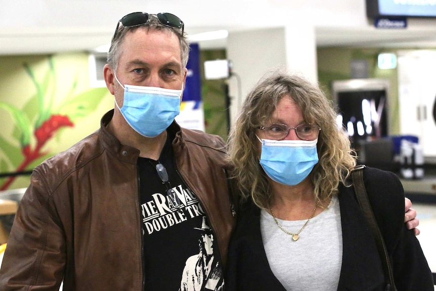 A man and a woman stand posing for a photo in an airport terminal wearing face masks, with the man's arm around the woman.
