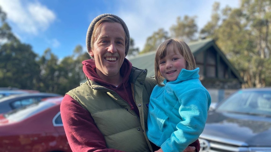 A man smiles and holds a small child in a car park with the wilderness in the background