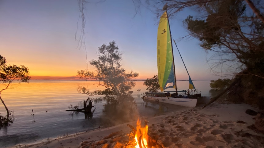 Hobie Cat pulled up on the beach with a small fire beside the water at sunset.