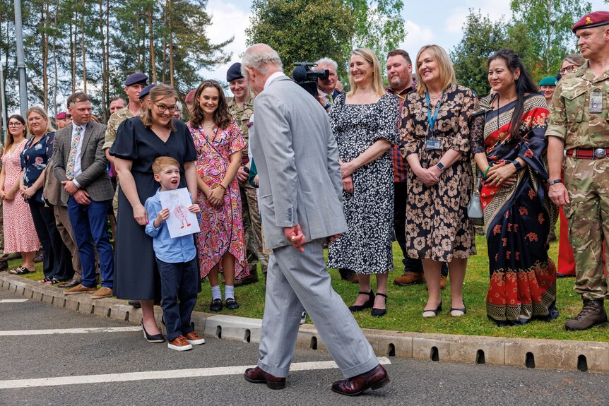 King Charles in a grey suit walking down a road lined with people and a little boy showing him a drawing.