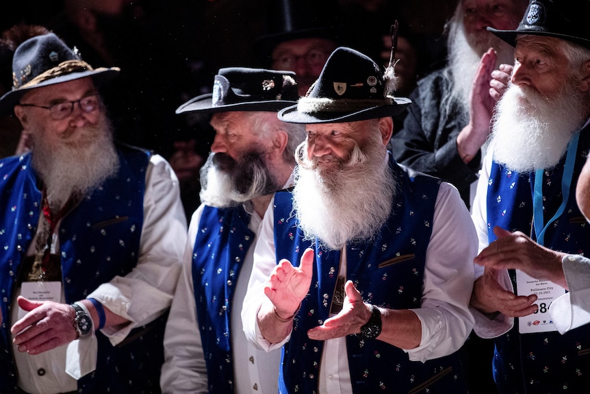 Men in white tops, blue vests and long white beards dancing and clapping