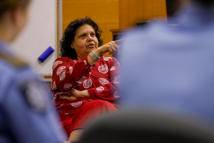 Woman in a red top points while explaining something with police recruits in blue uniform slightly blurred in foreground
