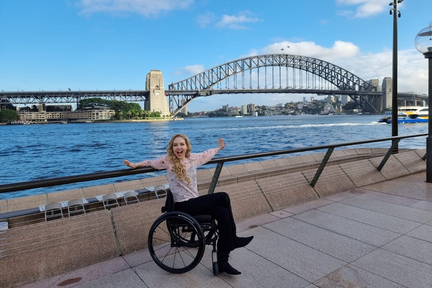 White woman with long blonde hair sitting in a wheelchair with arms outstretched in front of Sydney Harbor Bridge. 