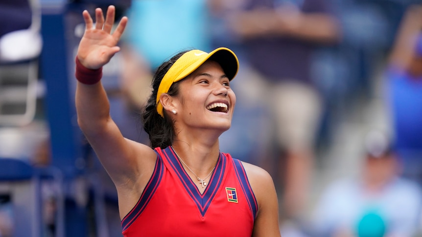 Emma Răducanu, wearing a yellow visor and red tennis singlet, smiles and waves to fans after a win at the US Open tennis.