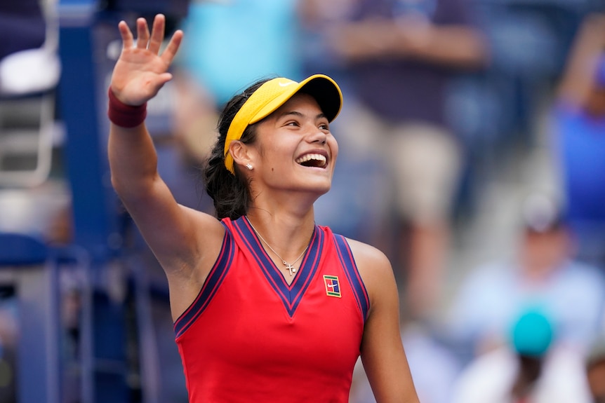 Emma Răducanu, wearing a yellow visor and red tennis singlet, smiles and waves to fans after a win at the US Open tennis.