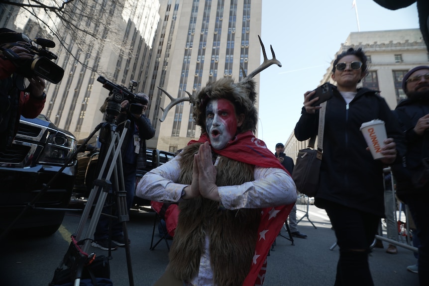 A man wearing a Trump flag as a cape, a woollen vest and antlers makes a prayer symbol