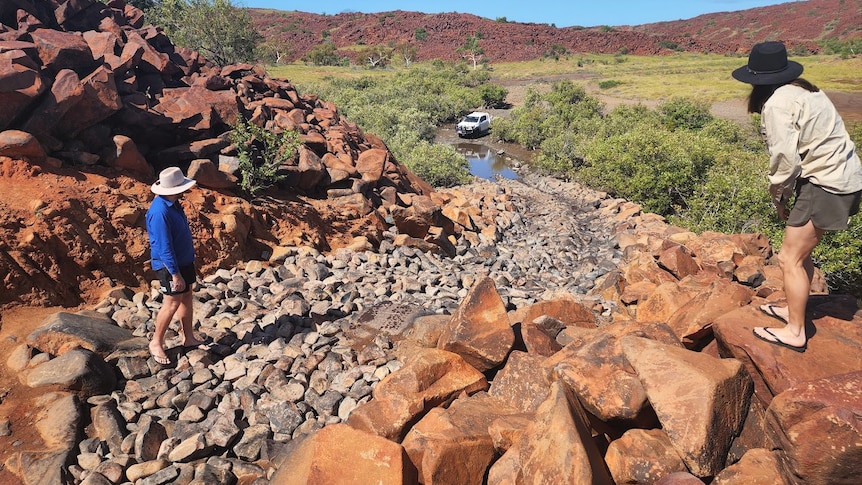 Two people watch as a four-wheel-drive starts to climb a rocky hill.