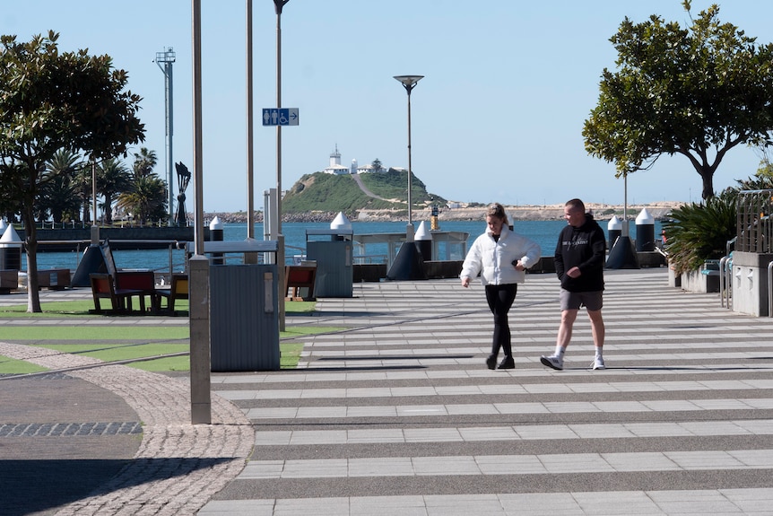 Two people walking along the promenade in Newcastle with the ocean in the background.