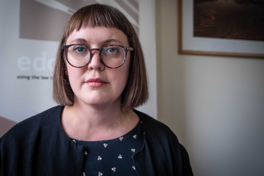 A woman with short brown hair and round glasses looks directly into the camera.