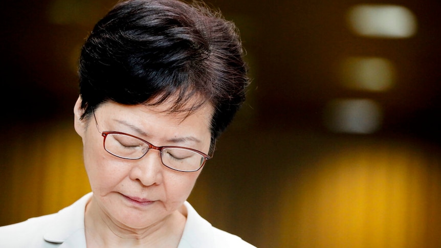 Against a soft background, a close up of Carrie Lam shows her with her eyes closed while wearing a cream suit.
