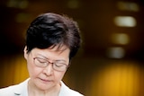 Against a soft background, a close up of Carrie Lam shows her with her eyes closed while wearing a cream suit.