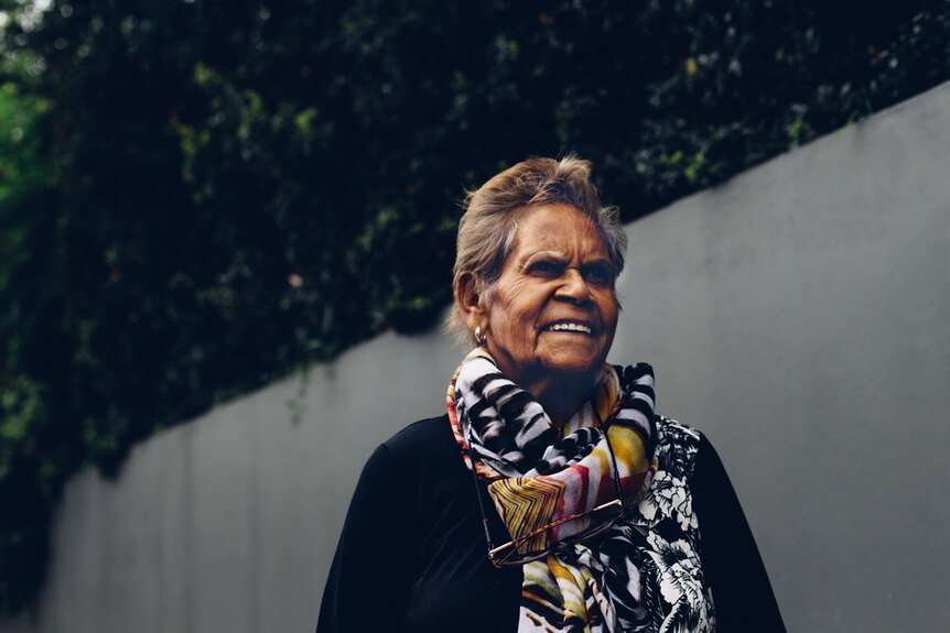 Portrait of an Indigenous woman wearing a colourful scarf and standing in front of a high concrete fence.