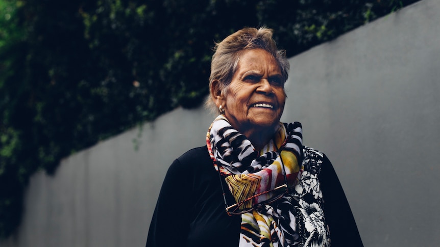 Portrait of an Indigenous woman wearing a colourful scarf and standing in front of a high concrete fence.