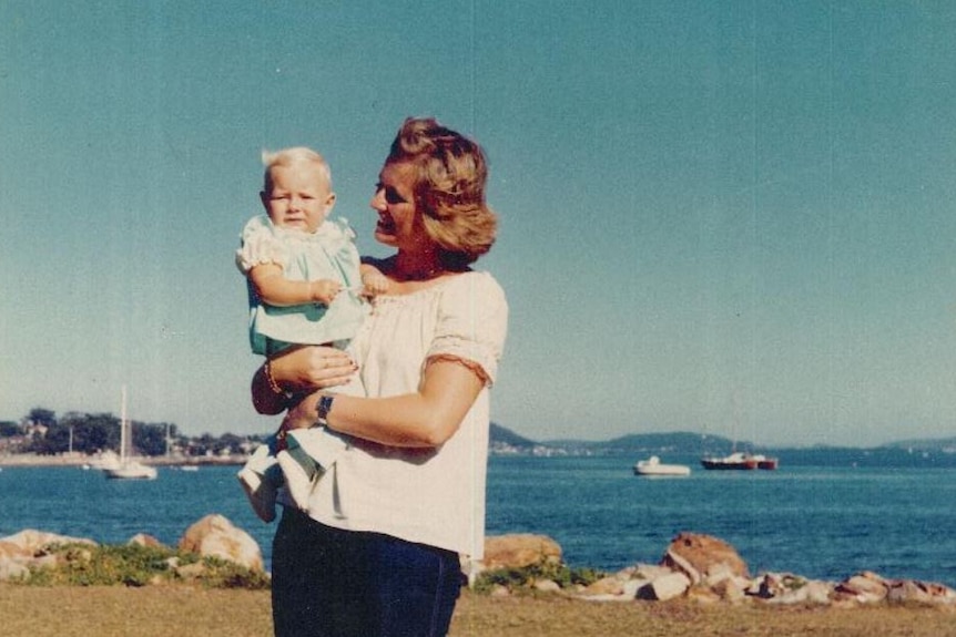 Smiling woman holds a blonde baby in a blue dress with the ocean in the background.