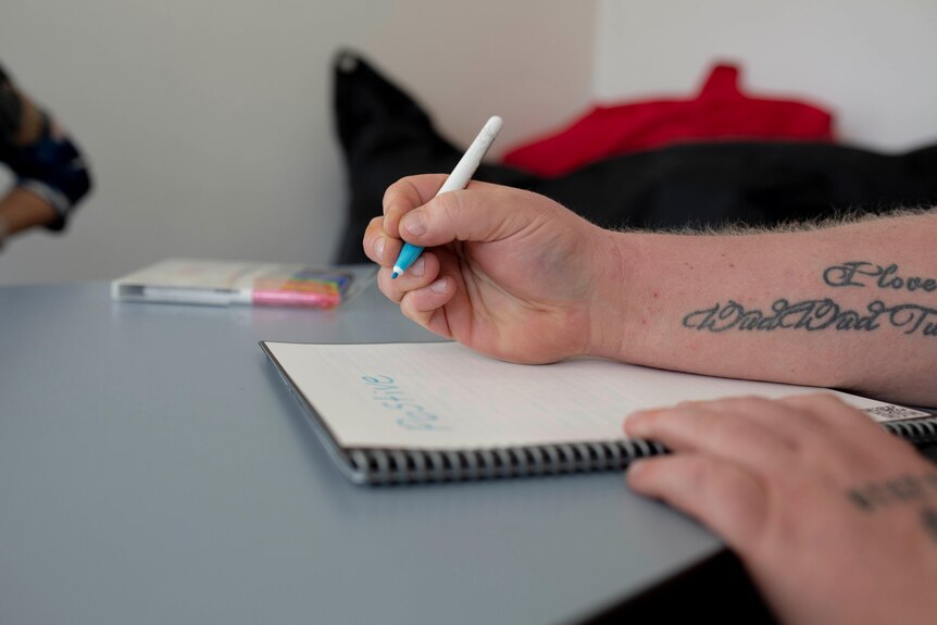A man's hand is visible as he writes in a notebook with a coloured pen.