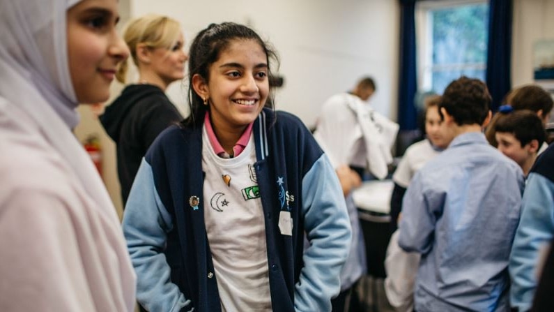 Two girls in school uniforms stand in front of other blurred students smiling and looking to the side.