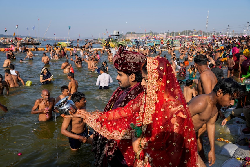 An Indian couple in formal traditional dress offer water from a silver urn to a river full of people swimming.