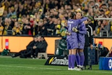 Two soccer players wearing purple hug on the sidelines of a game with a big crowd in the background