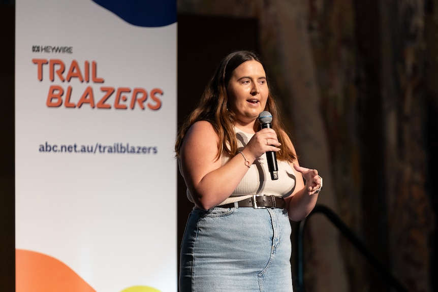 A young woman is pictured speaking into a microphone, behind her there's a sign that says 'ABC Heywire, Trailblazers'.