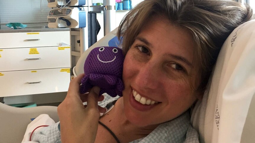 Ms White smiles, holding a purple jellyfish toy to her face while laying on a hospital bed.