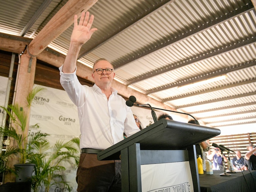 Man in white shirt standing behind a podium waves to the camera