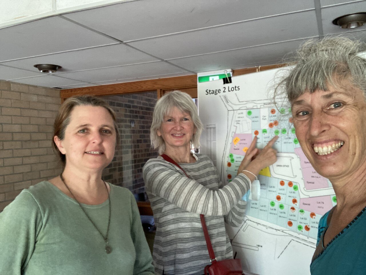 Three women smile in front of a board showing a development plan. 