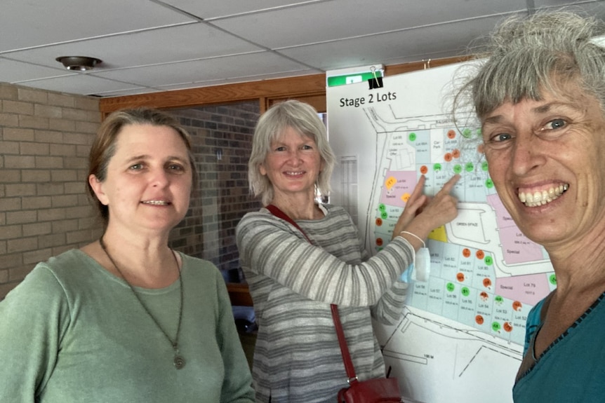 Three women smile in front of a board showing a development plan. 