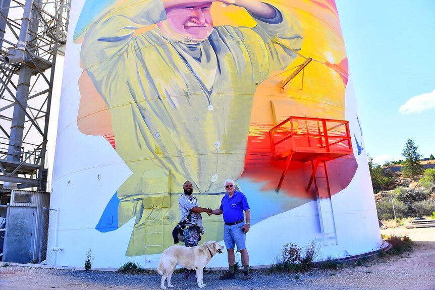 A giant silo painted with a picture of a man looking out across the horizon. There are two men shaking hands and a dog.