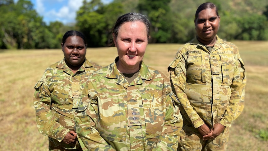 Three women in army fatigues pose for a photograph in a grassy clearing.