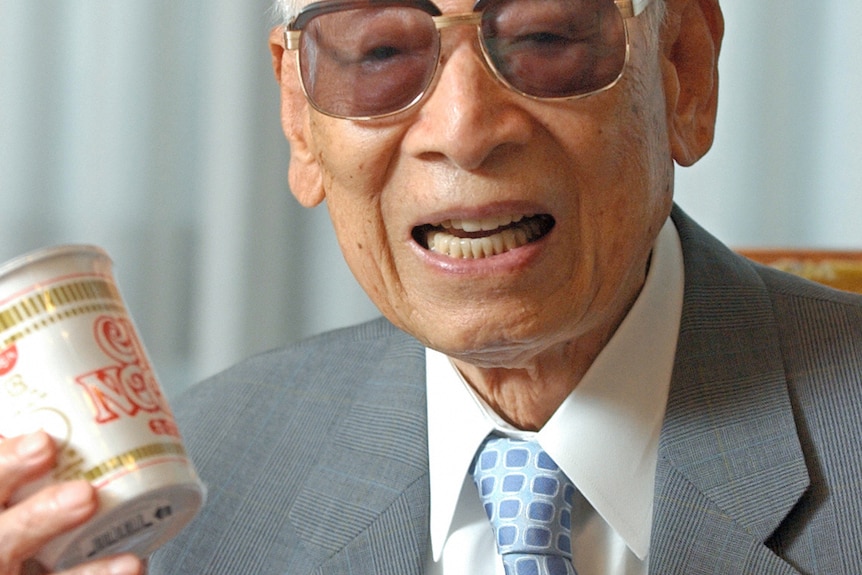 Picture of and elderly Momofuku Ando holding a noodle cup wearing shaded spectacles, grey suit jacket and blue checkered tie.