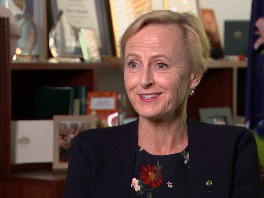 A woman with short blonde hair wearing a black blazer mid-sentence and smiling