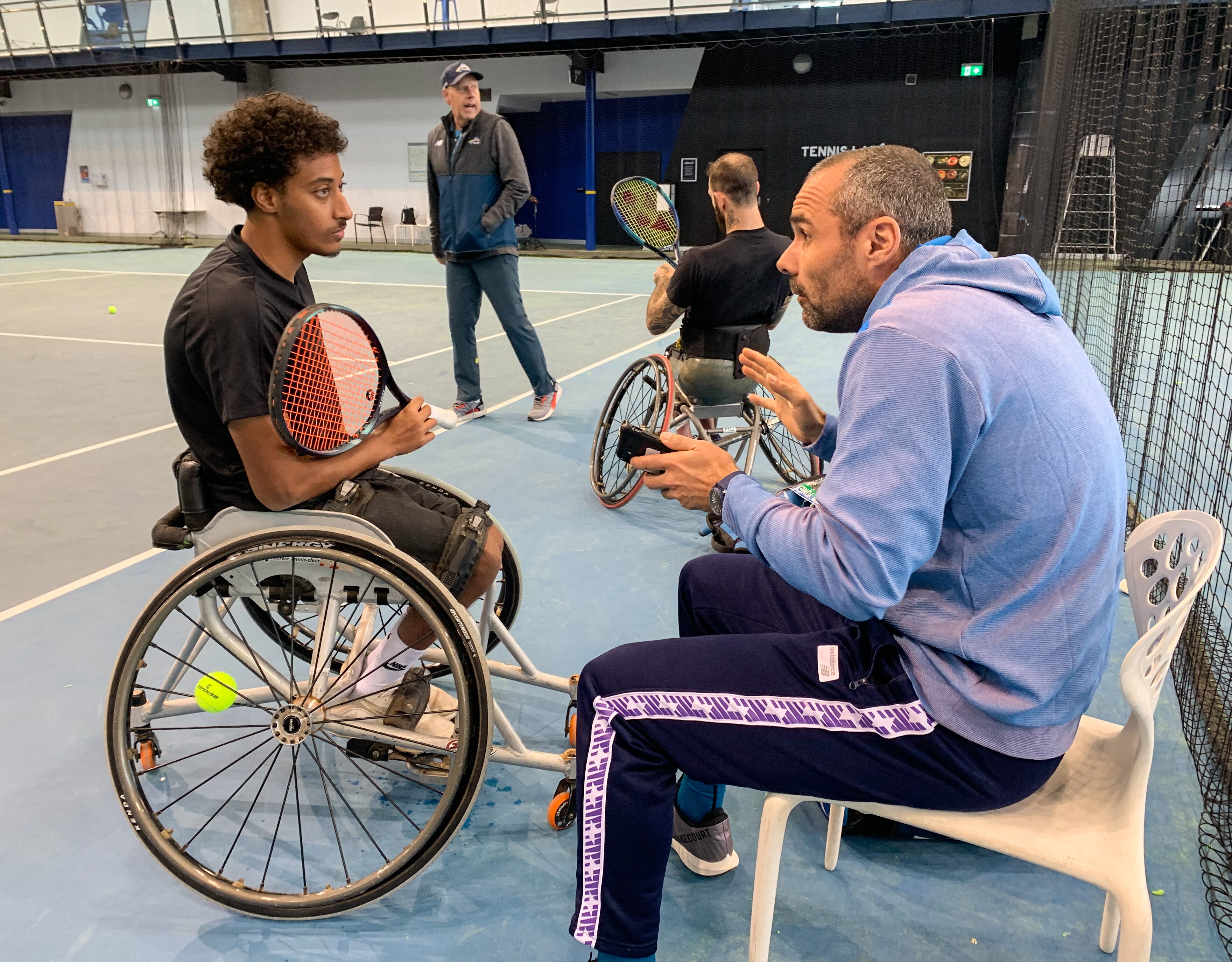 A wheelchair tennis player sits at courtside holding his racquet, listening to his coach, who is gesturing with his hands.