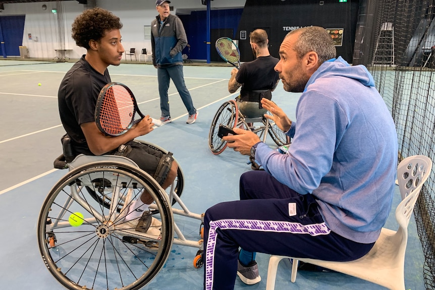 A wheelchair tennis player sits at courtside holding his racquet, listening to his coach, who is gesturing with his hands.