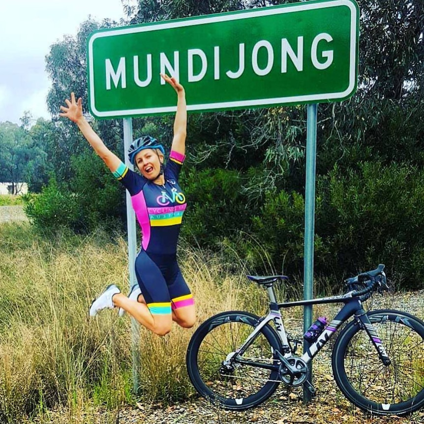 A women wearing cycling clothes jumps in the air, next to her bike and in front of a sign reading "Mundijong".