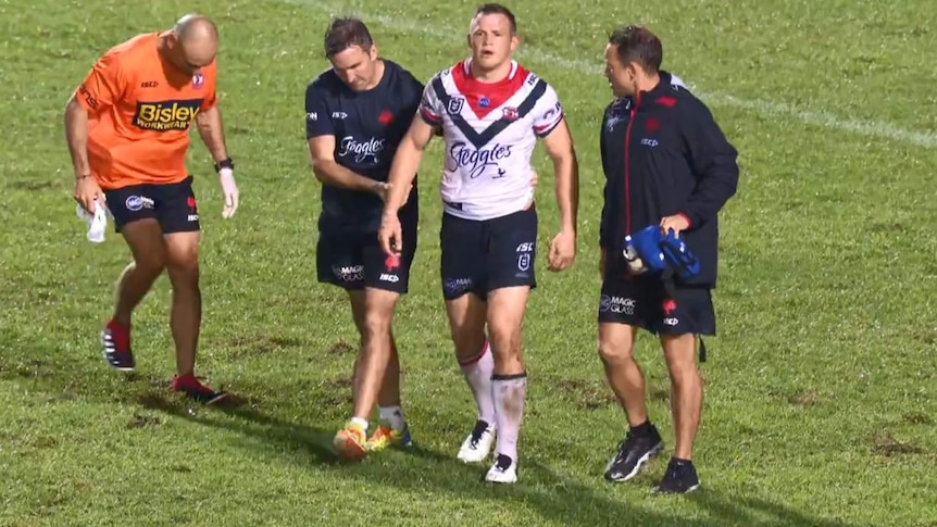 Brett Morris limps off the field with two men in black coats either side and a man in orange stamping on the grass to the left