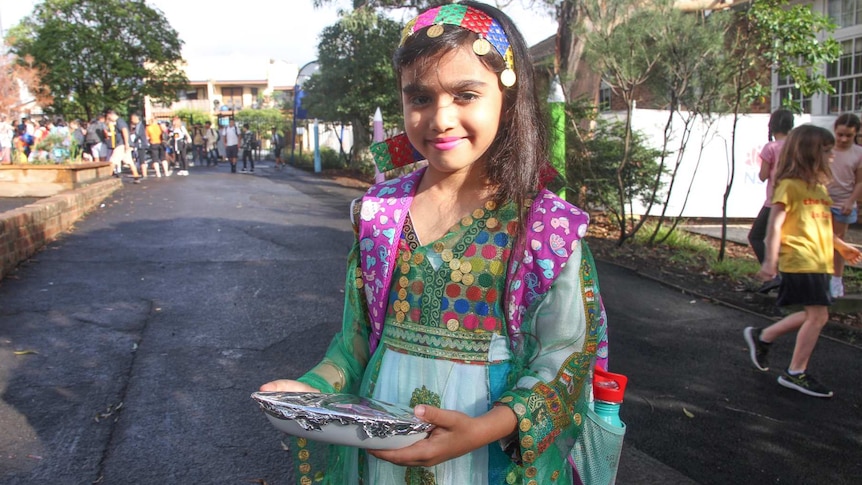 A girl, wearing colourful traditional clothes, carrying a plate of food outside a school